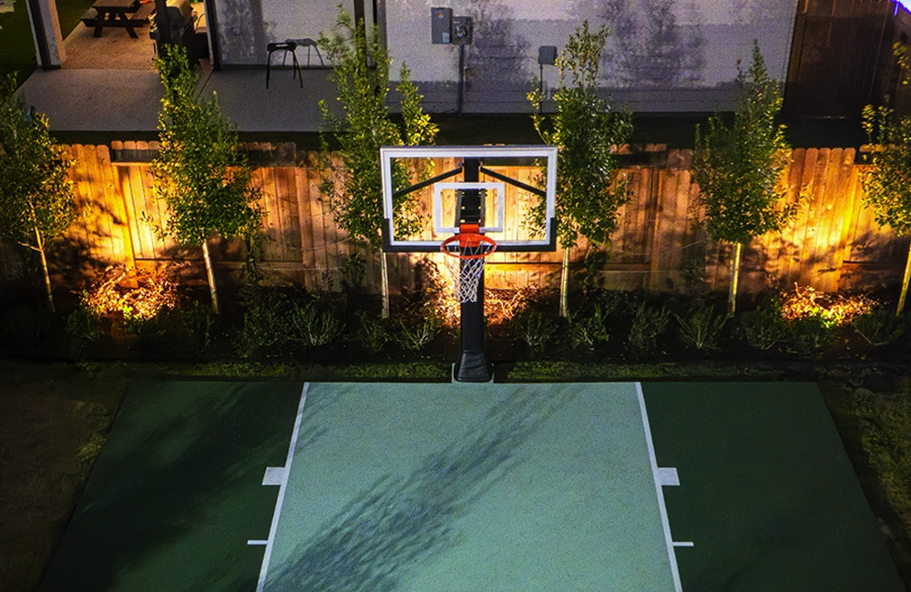 Residential basketball court and landscape illuminated with tree uplighting and warm outdoor lighting accents at night.