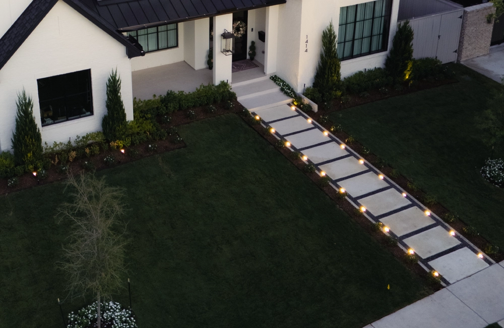 Aerial evening view of illuminated walkway and driveway lighting guiding entry through a modern residential landscape.